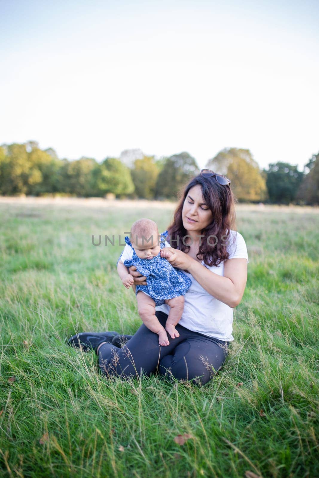 Happy woman sitting on grass and holding her cute baby with trees as background. Mother and daughter relaxing in park under clear sky. Young family having fun outdoors