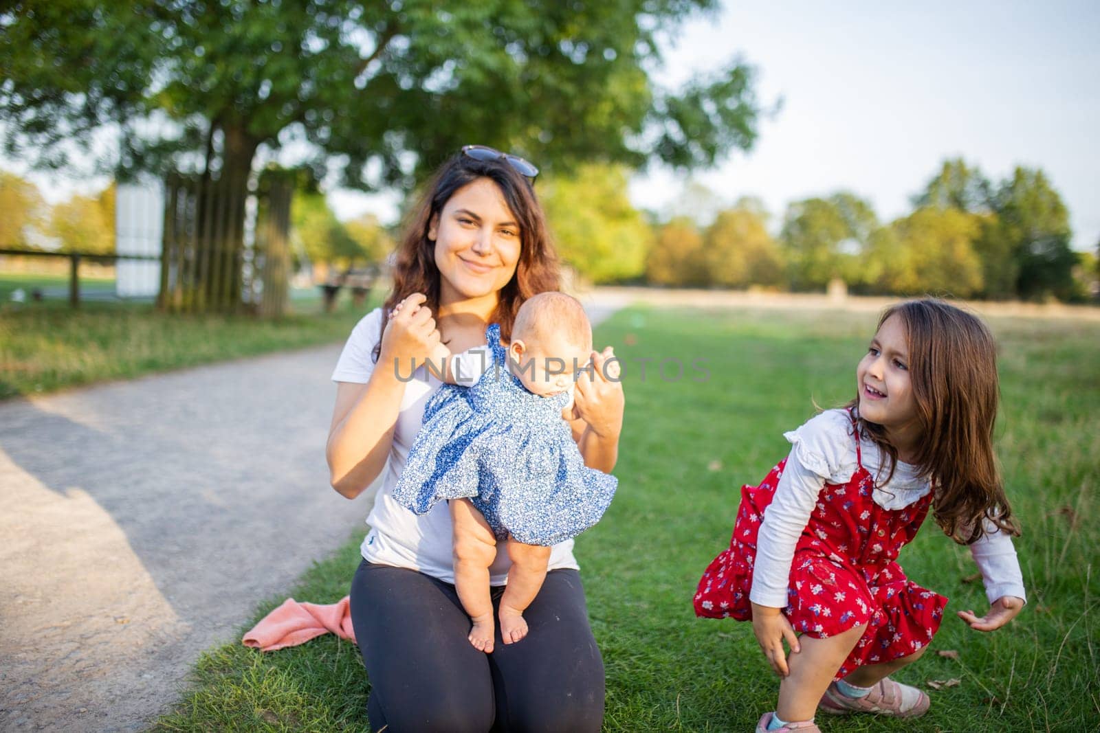 Happy woman, baby, and little girl sitting on grass with trees as background. Smiling mother playing with adorable daughters in park. Young family having fun outdoors