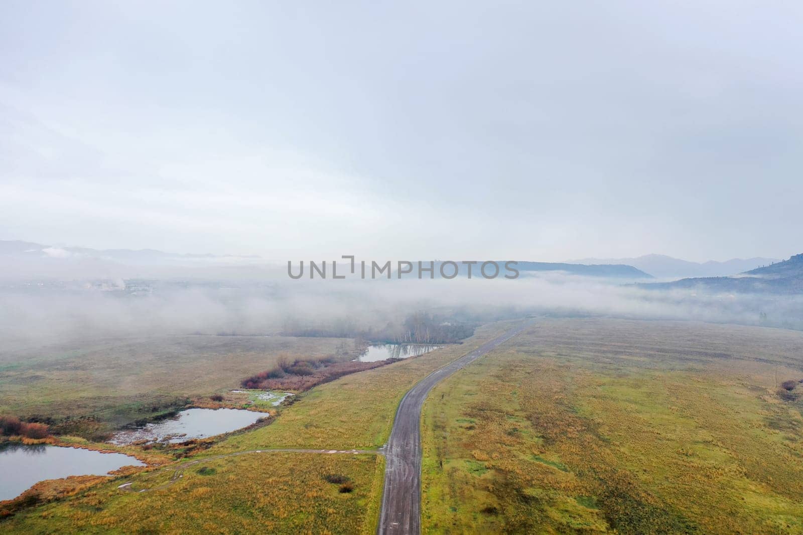 Road in the middle of open field and under cloudy sky with mountains as background. Aerial view of paved road and marsh on empty grassland. Foggy landscapes
