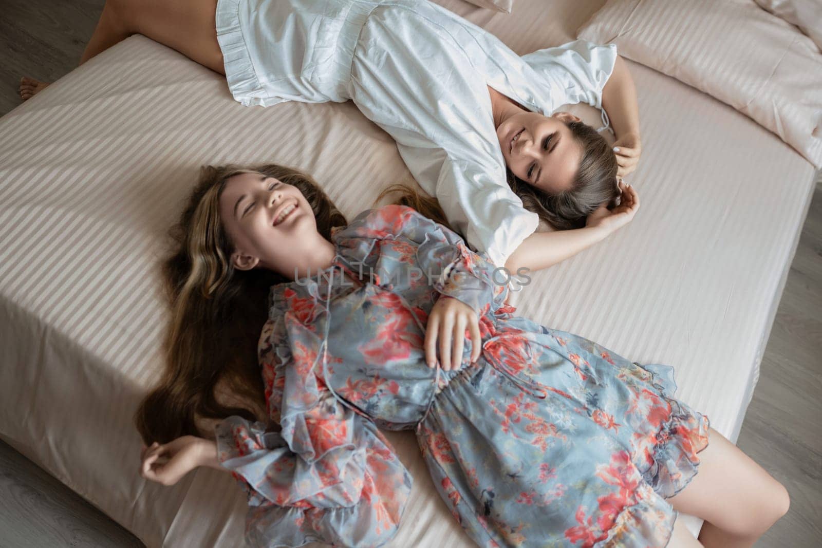 Two women lie laughing on a bed. One wears a floral dress, the other a white robe. Relaxed, joyful scene.