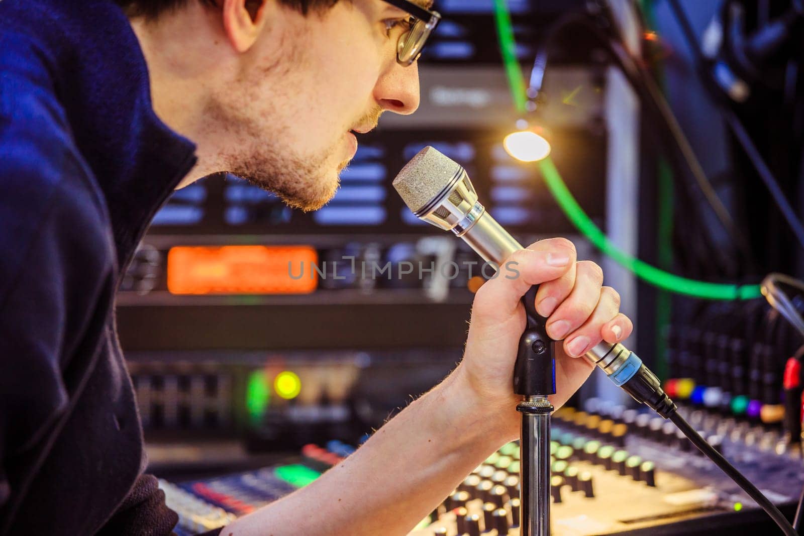 Young man is sitting in the recording studio and talks into the microphone, buttons and equipment in the blurry background