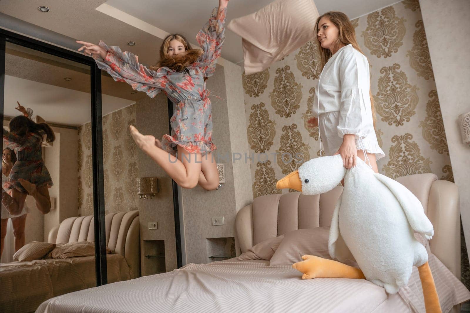 Two women in a bedroom. One joyfully leaps the other holds a large stuffed bird on a bed.