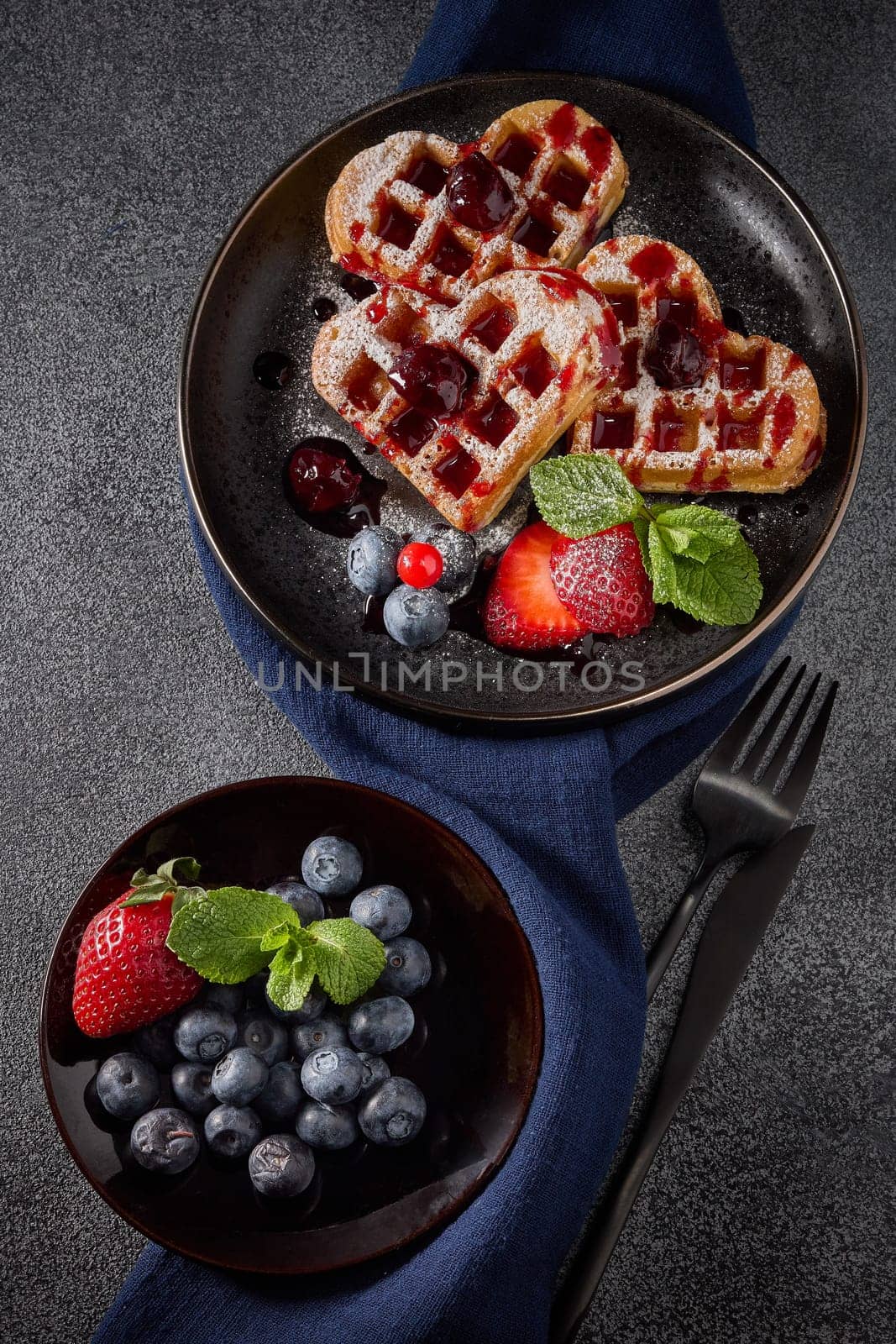 Delicious waffles heart shape with  powdered sugar and berries on ceramic black plate and saucer with berries on grey background. Top view. Sweet meal. Dessert. Serving food