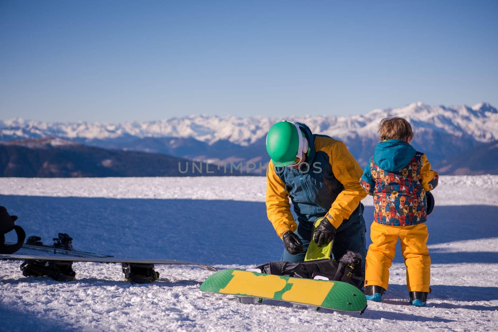 young happy father preparing his little son for the first time on a snowboard during sunny winter day at beautiful  ski resort