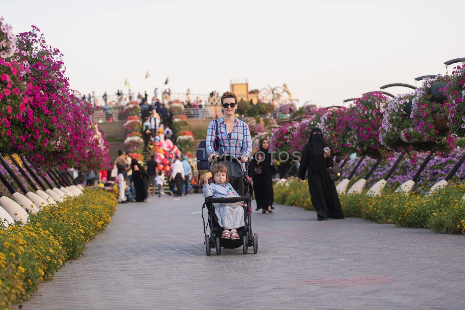 young mother and cute little girl in a baby stroller enjoying a beautiful day in the flower garden