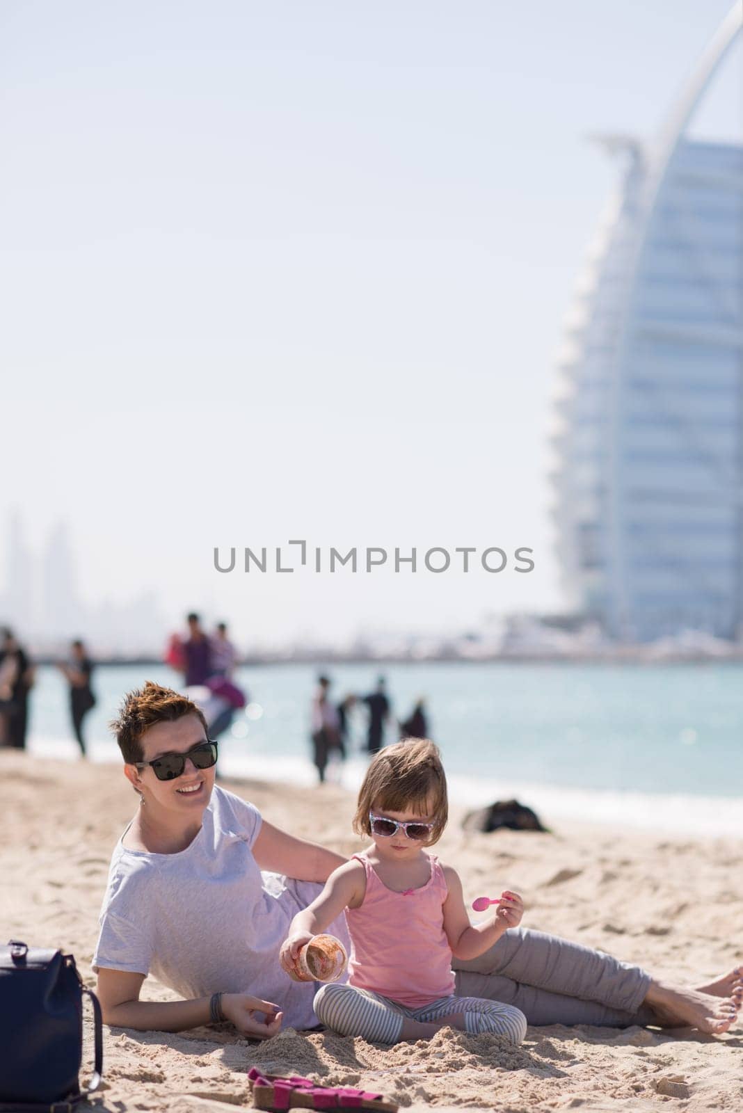 cool young mom and her little girl playing with a bucket full of sand wearing sunglasses and relaxing on the beach on a sunny day
