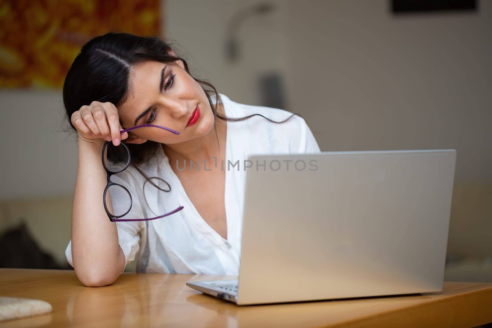 Tired woman supporting her head with hand and removing glasses while working from home. Fatigue caused by executing a job on remote device. Exhausted person sitting hopeless behind a laptop.