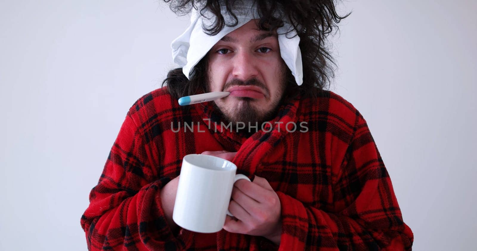 young Man with flu and fever wrapped holding cup of healing tea isolated over white
