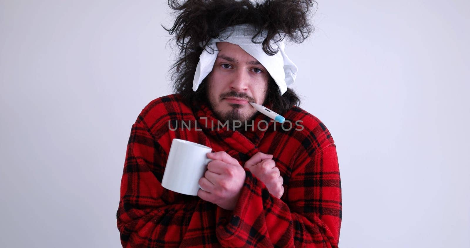 young Man with flu and fever wrapped holding cup of healing tea isolated over white