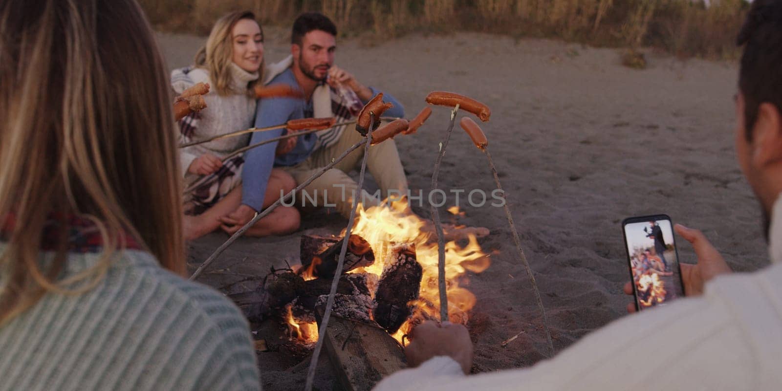 Group of young friends sitting by the fire late at night, grilling sausages and drinking beer, talking and having fun