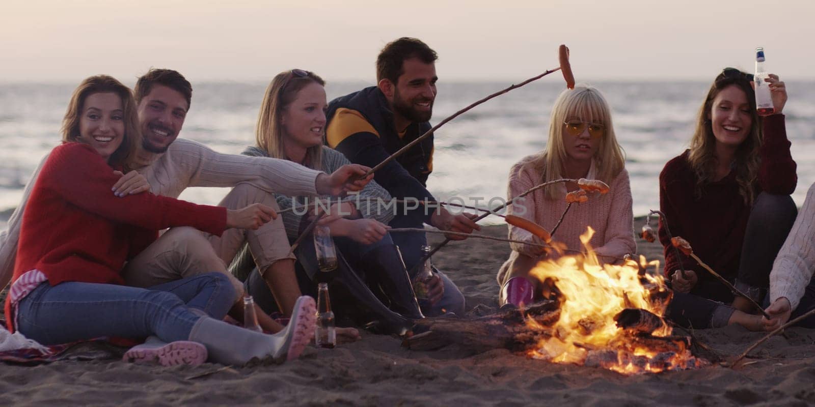 Group of young friends sitting by the fire late at night, grilling sausages and drinking beer, talking and having fun