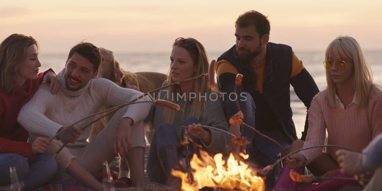 Group of young friends sitting by the fire late at night, grilling sausages and drinking beer, talking and having fun