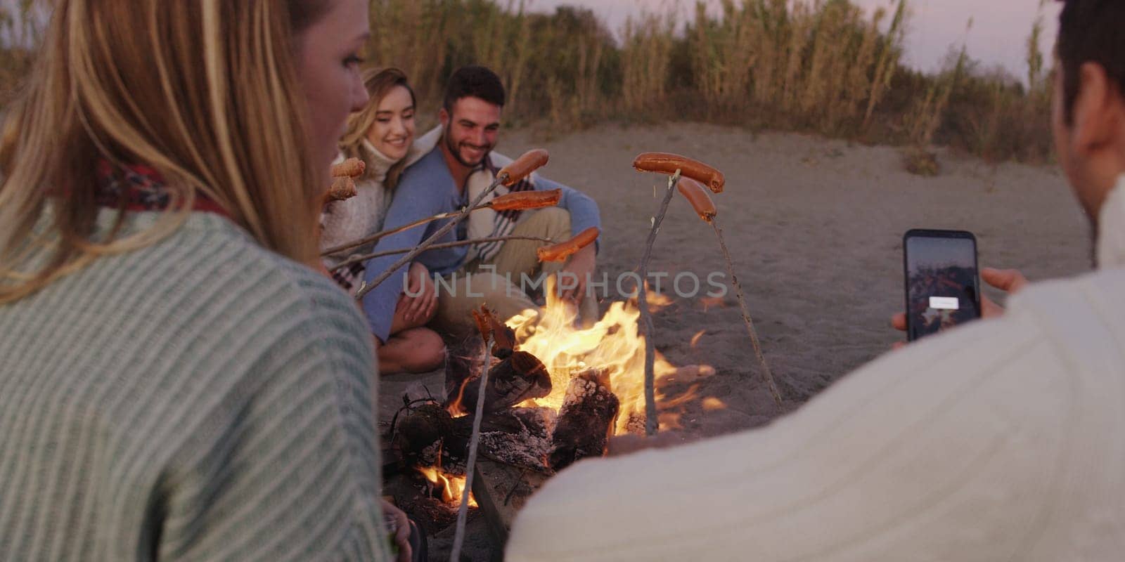 Group of young friends sitting by the fire late at night, grilling sausages and drinking beer, talking and having fun