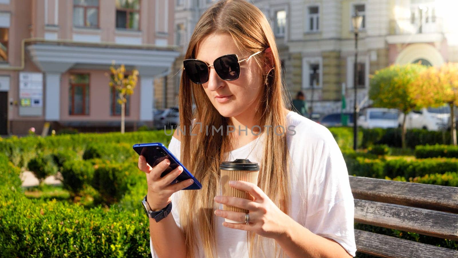 Portrait of female student drinking coffee and using smartphone in park.