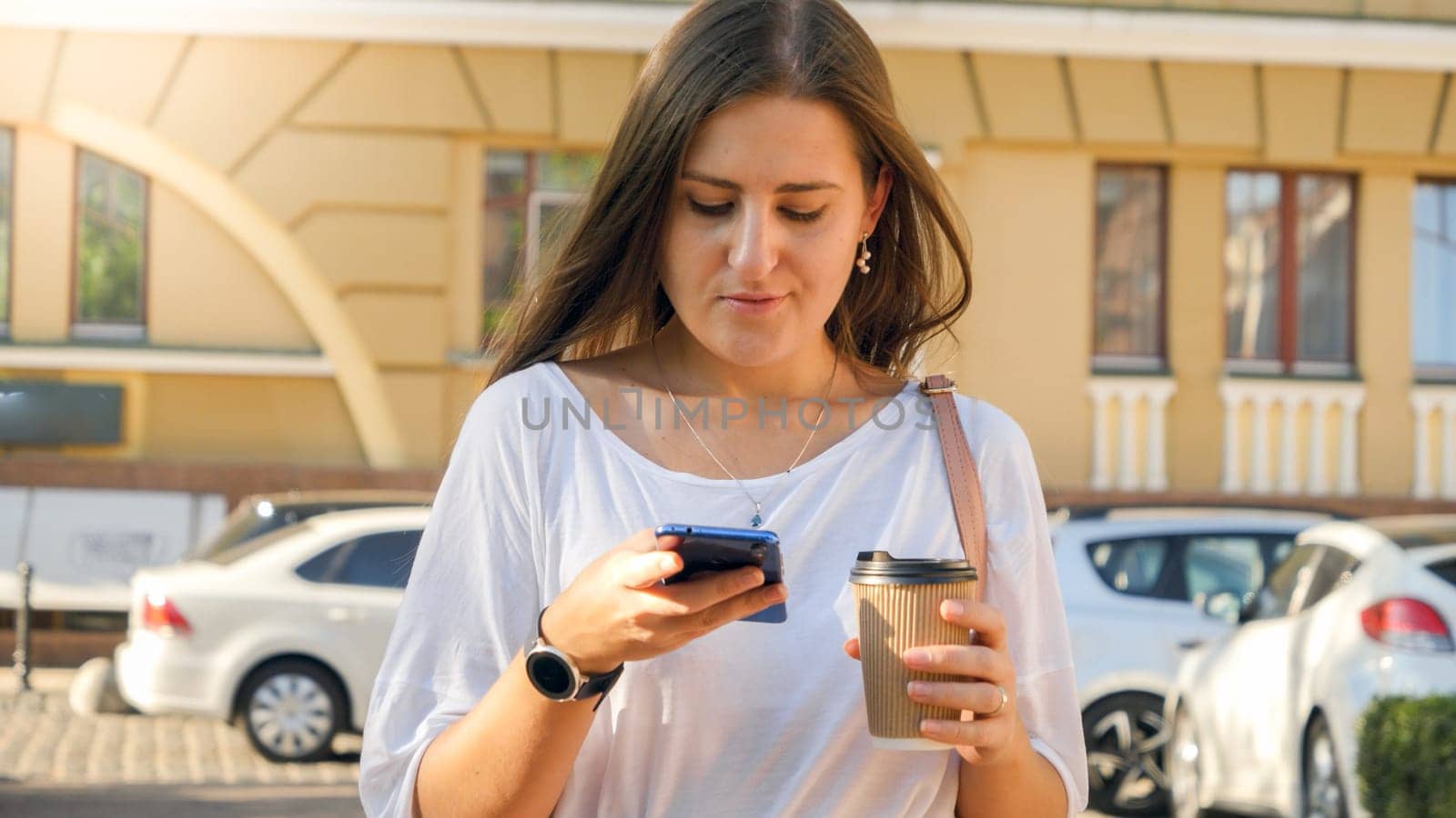 Portrait of beautiful female worker walking on city street with coffe cup and browsing smartphone.