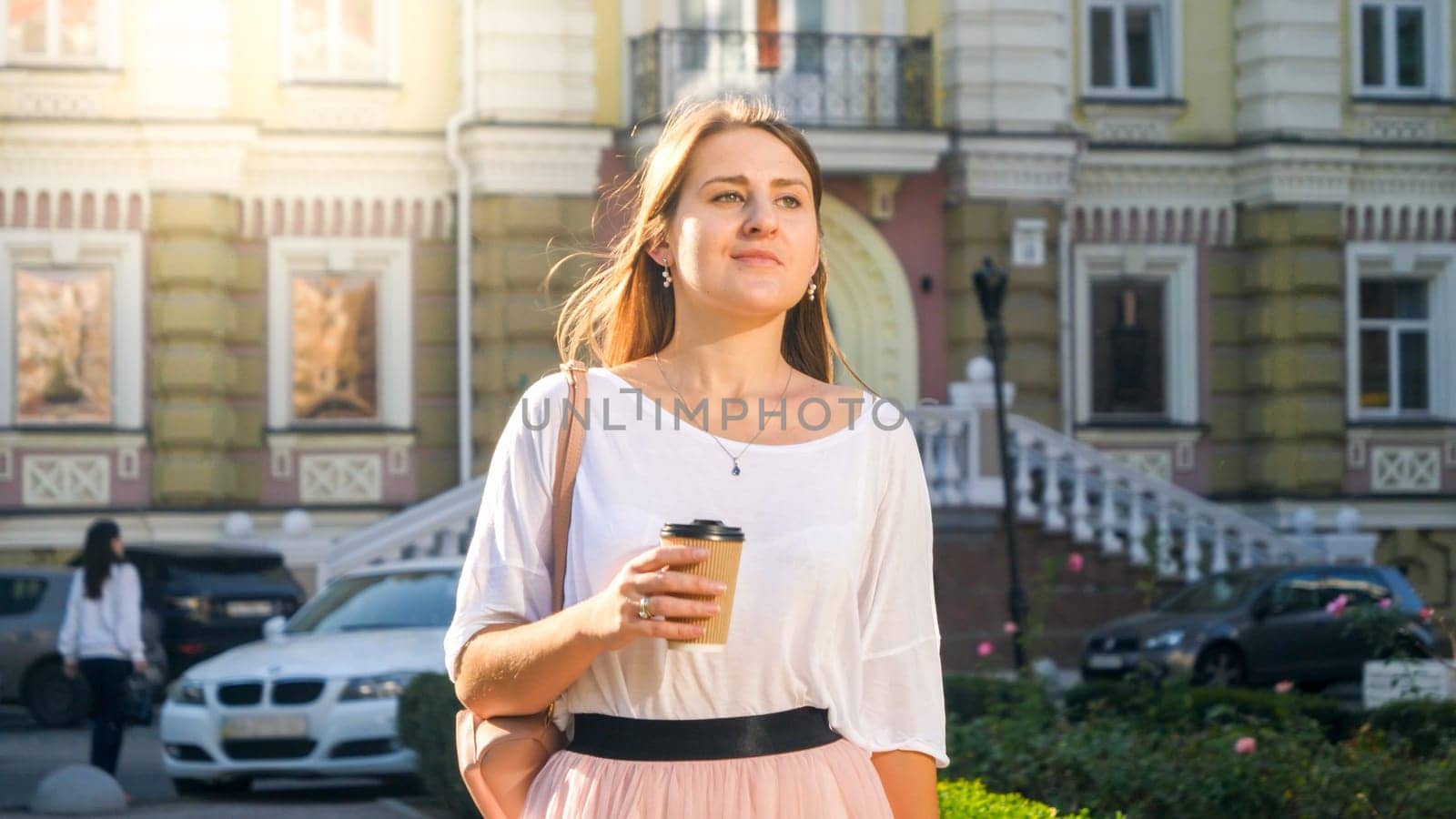 Portrait of stylish young woman walking on city street with coffee-to-go paper cup.