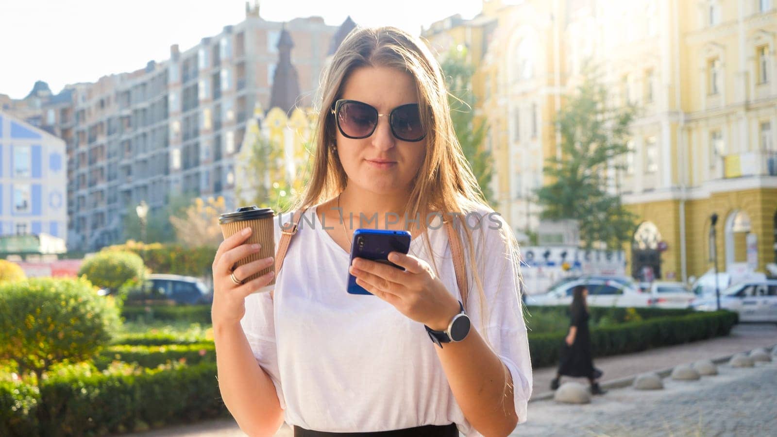 Portrait of beautiful young woman long hair holding coffee cup walking on street and typing message in social media on smartphone.