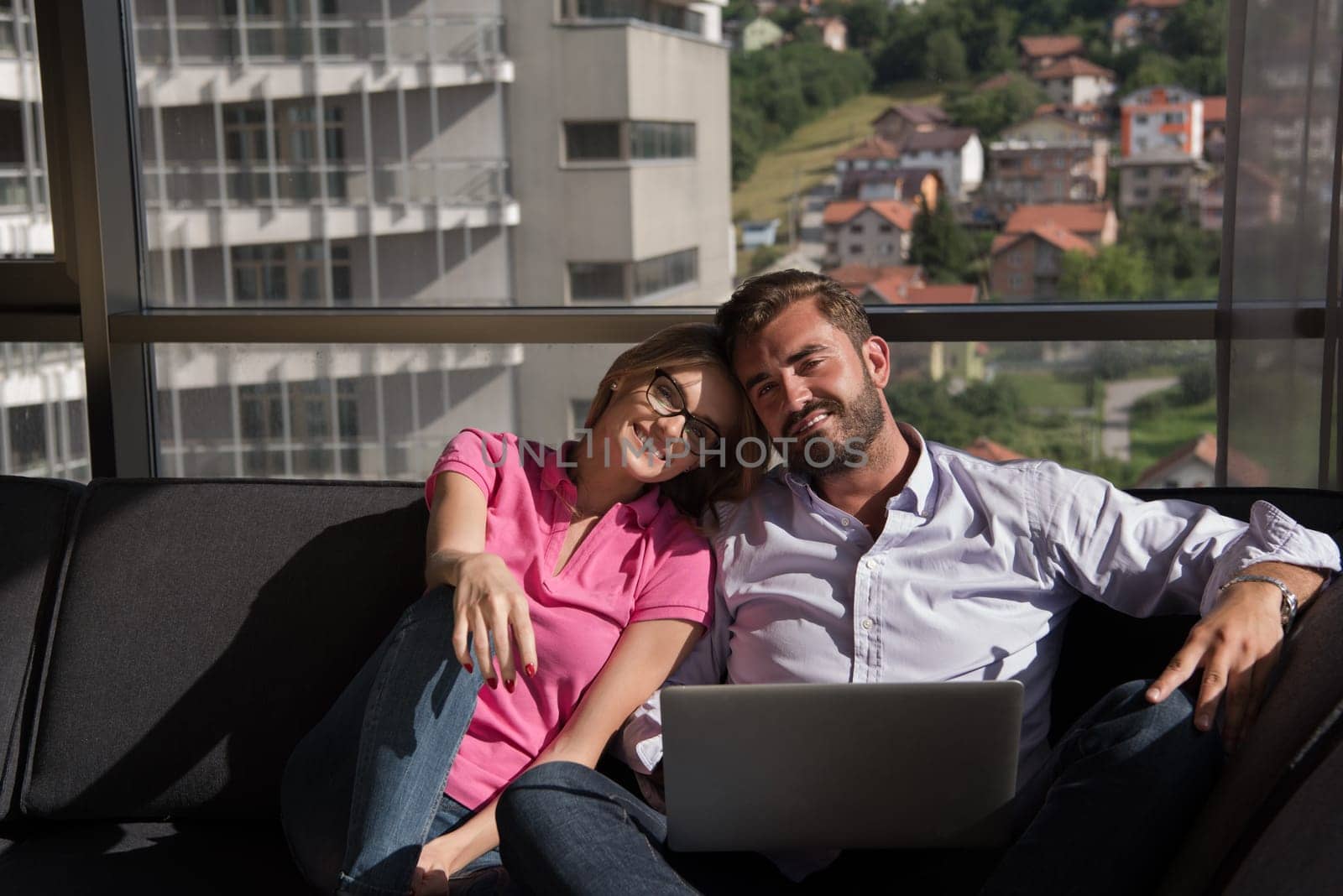 Young couple relaxing at  home using laptop computers reading in the living room near the window on the sofa couch.