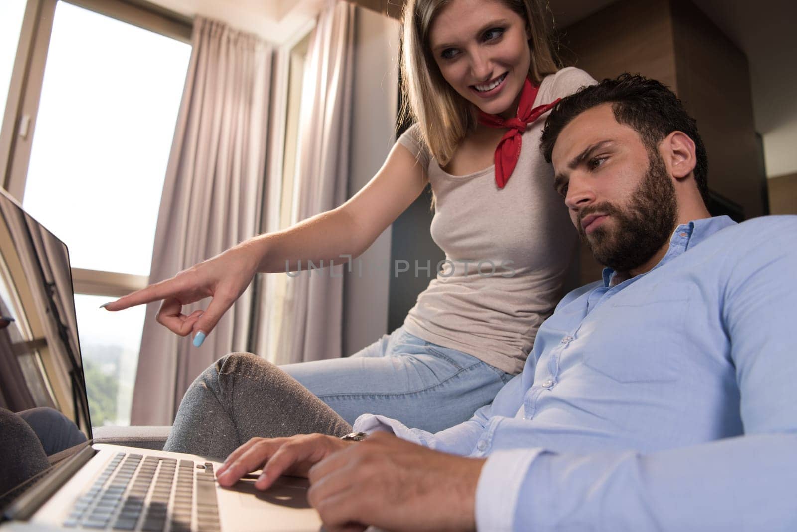 Young couple relaxing at  home using laptop computers reading in the living room near the window on the sofa couch.