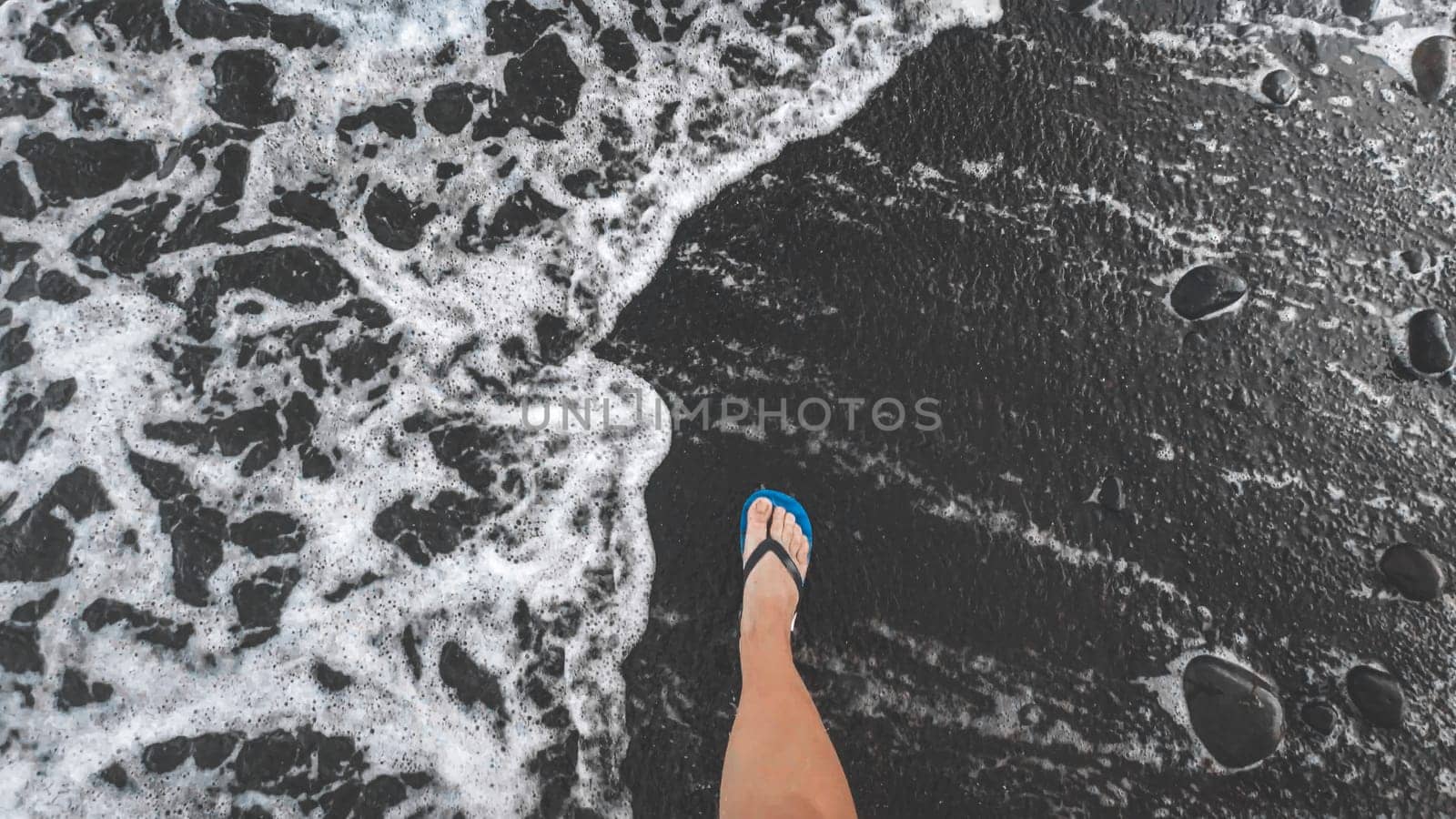 View from the top on male feet in flip flops walking on black volcanic sandy sea beach.