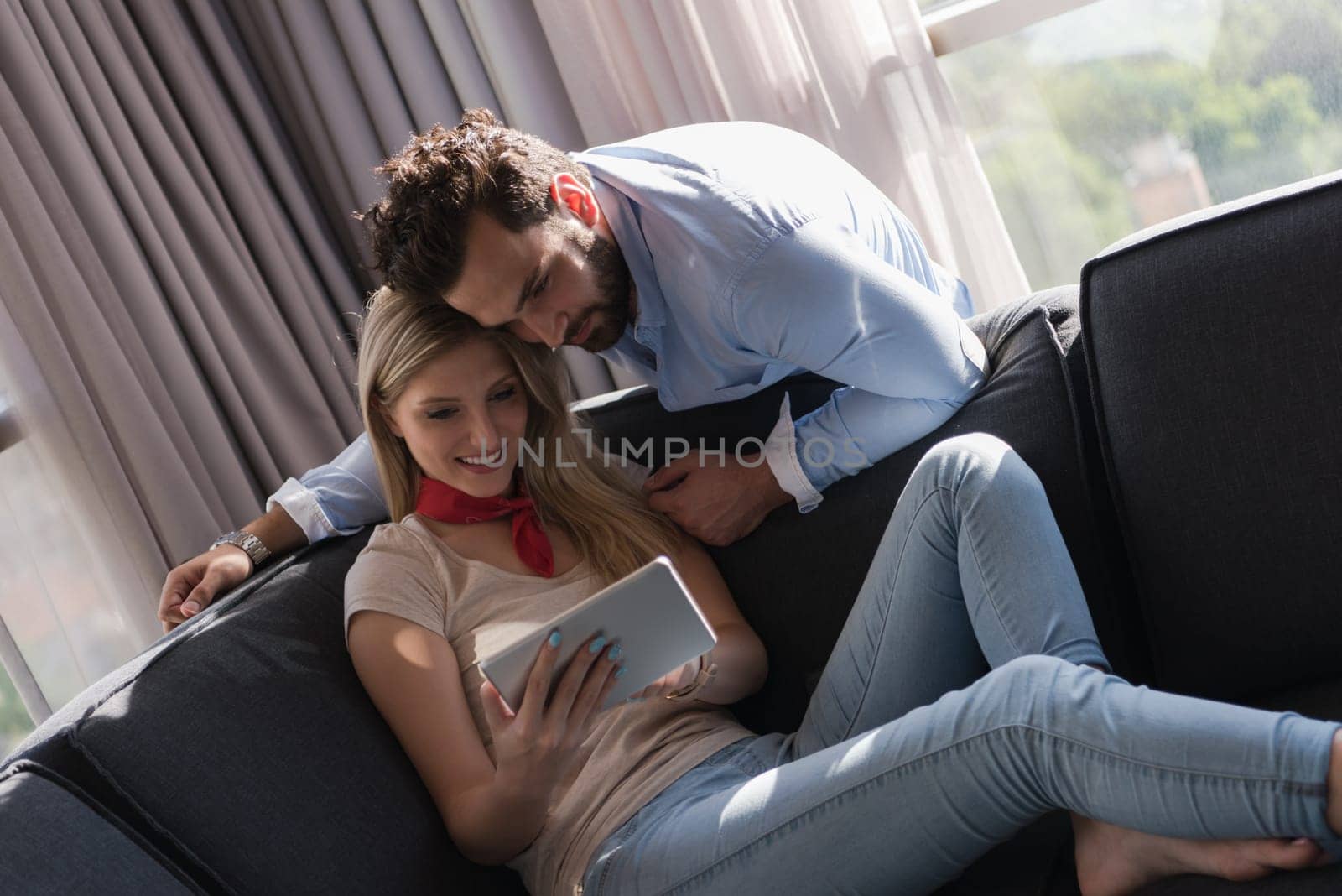 Young couple relaxing at  home using tablet computer reading in the living room near the window on the sofa couch.