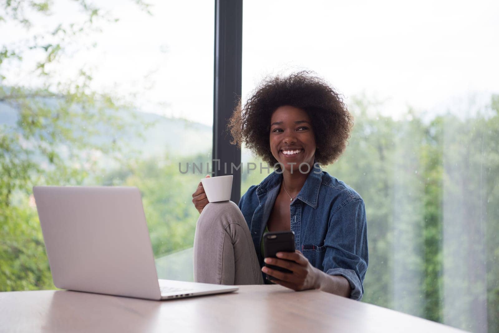Young african american woman smiling sitting near bright window while looking at open laptop computer on table and holding white mug in her luxury home