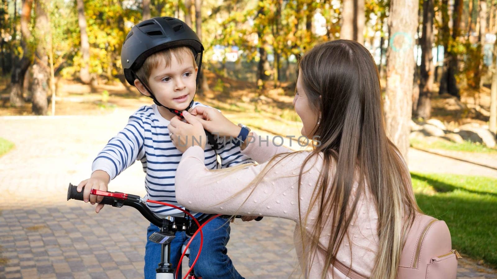 Portrait of beautiful young mother adjusting safety helmet on her son's head before riding a bicycle at park.