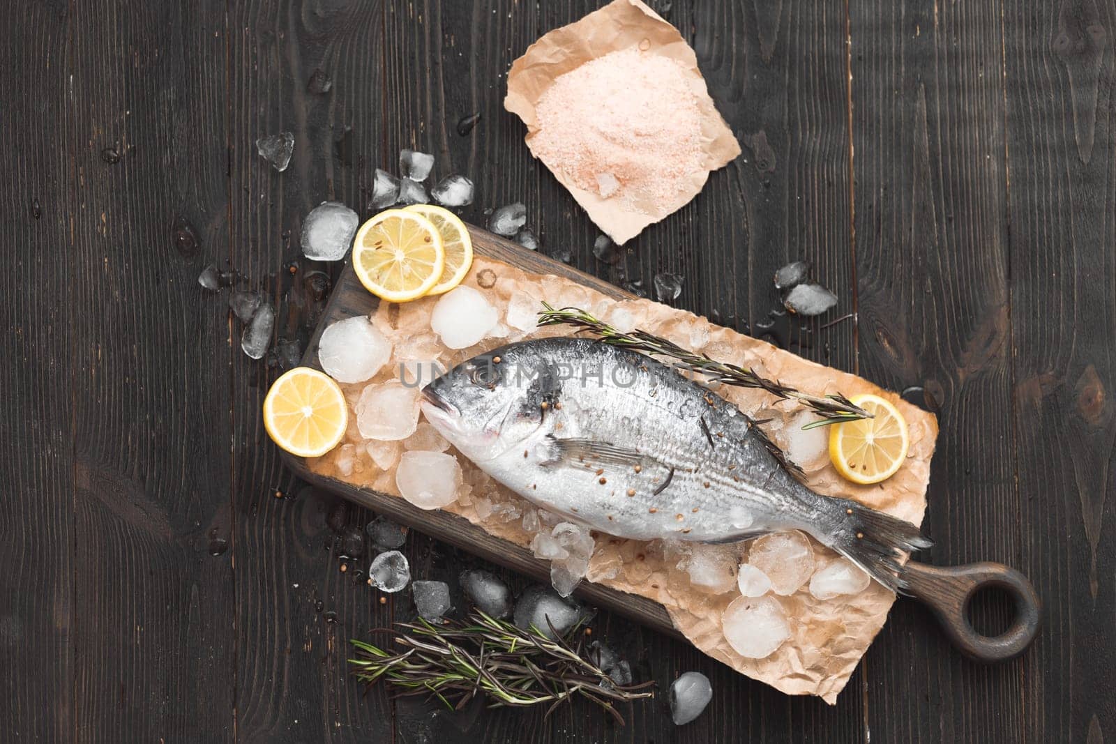 Raw dorada fish or gilt-head bream on ice with lemon slices, salt and rosemary over black wooden background, flat lay, top view