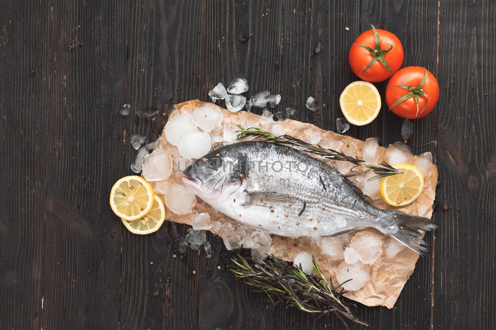 Raw dorada fish or gilt-head bream on ice with lemon slices and rosemary over black wooden background, flat lay, top view