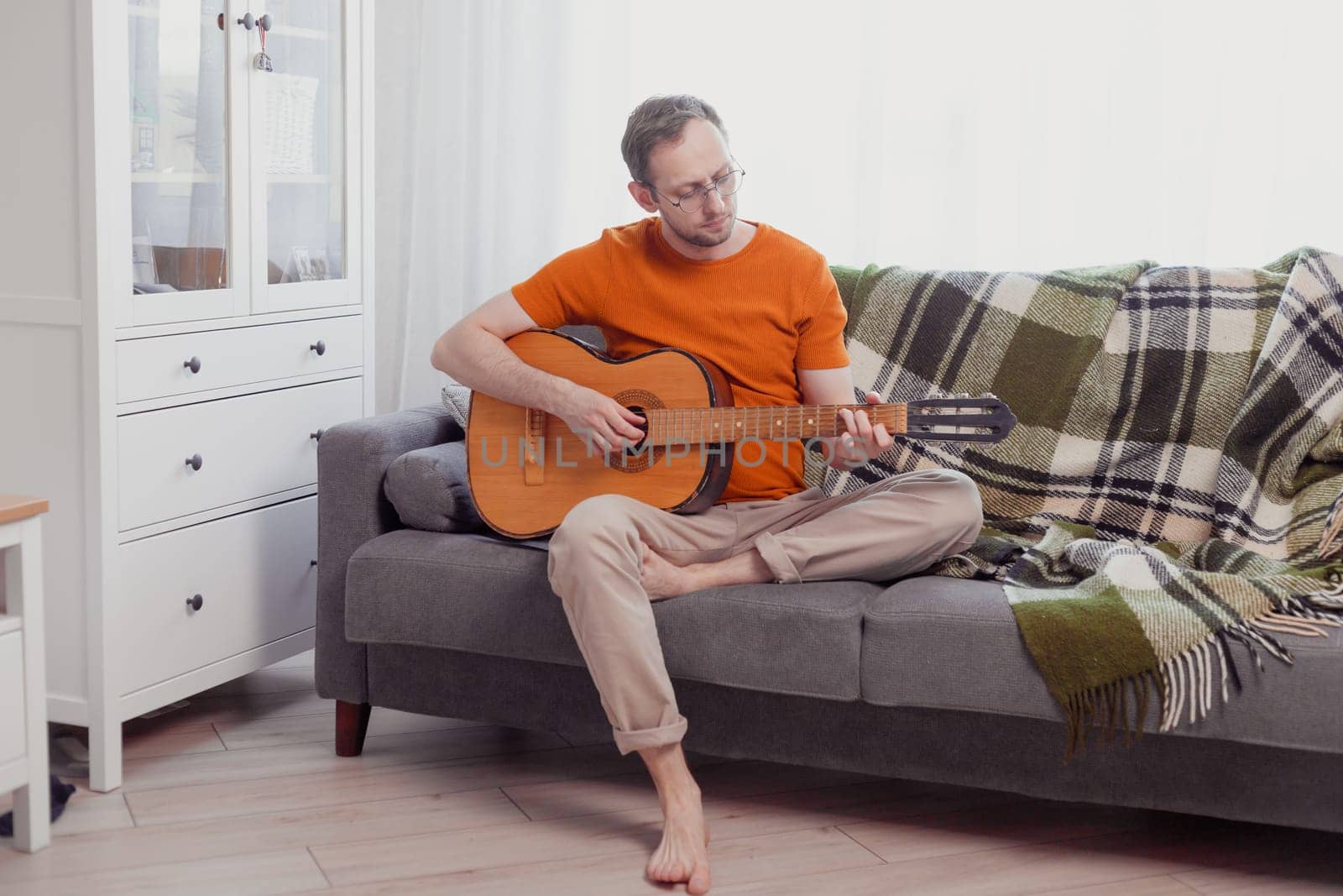 Young caucasian man playing the guitar sitting on couch at home. Practice music, lifestyle photo.