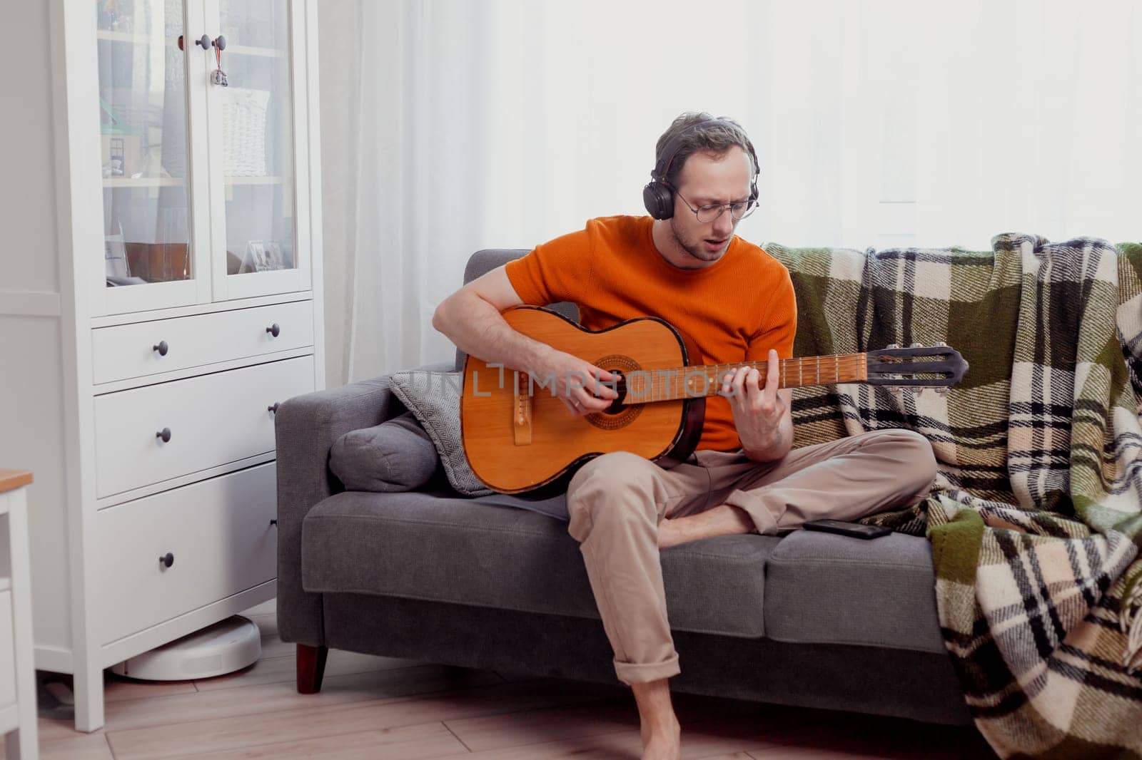 Young caucasian man in headphones playing the guitar sitting on couch at home. Practice music, lifestyle photo