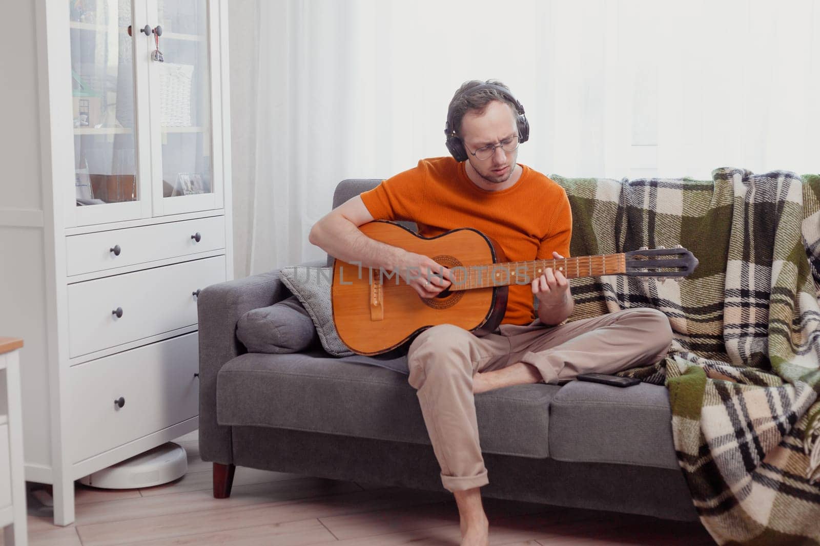 Young caucasian man in headphones playing the guitar sitting on couch at home. Practice music, lifestyle photo
