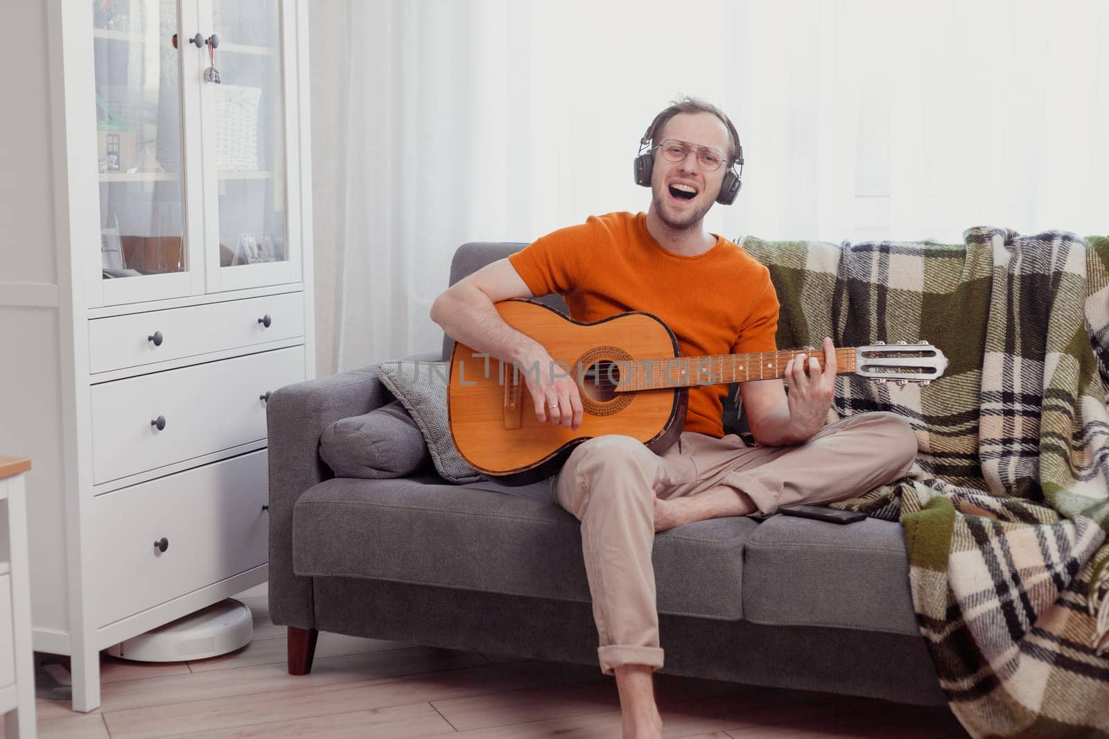 Young caucasian man in headphones playing the guitar and singing at home. Quarantine and hobbies concept. Practice music, lifestyle photo