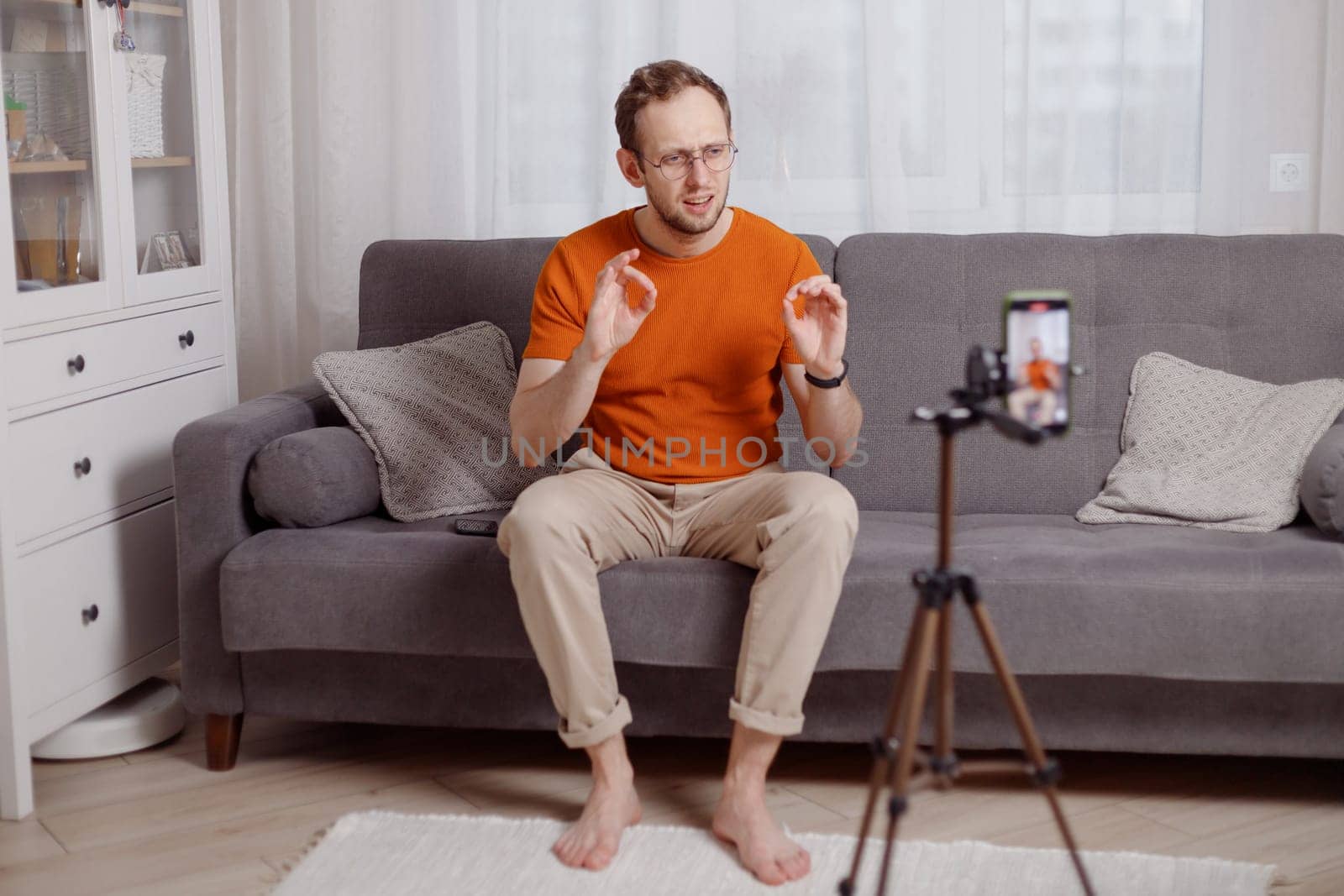 Hipster man blogger recording vlog on mobile phone sit on couch in living room and explain and gesturing with hands.
