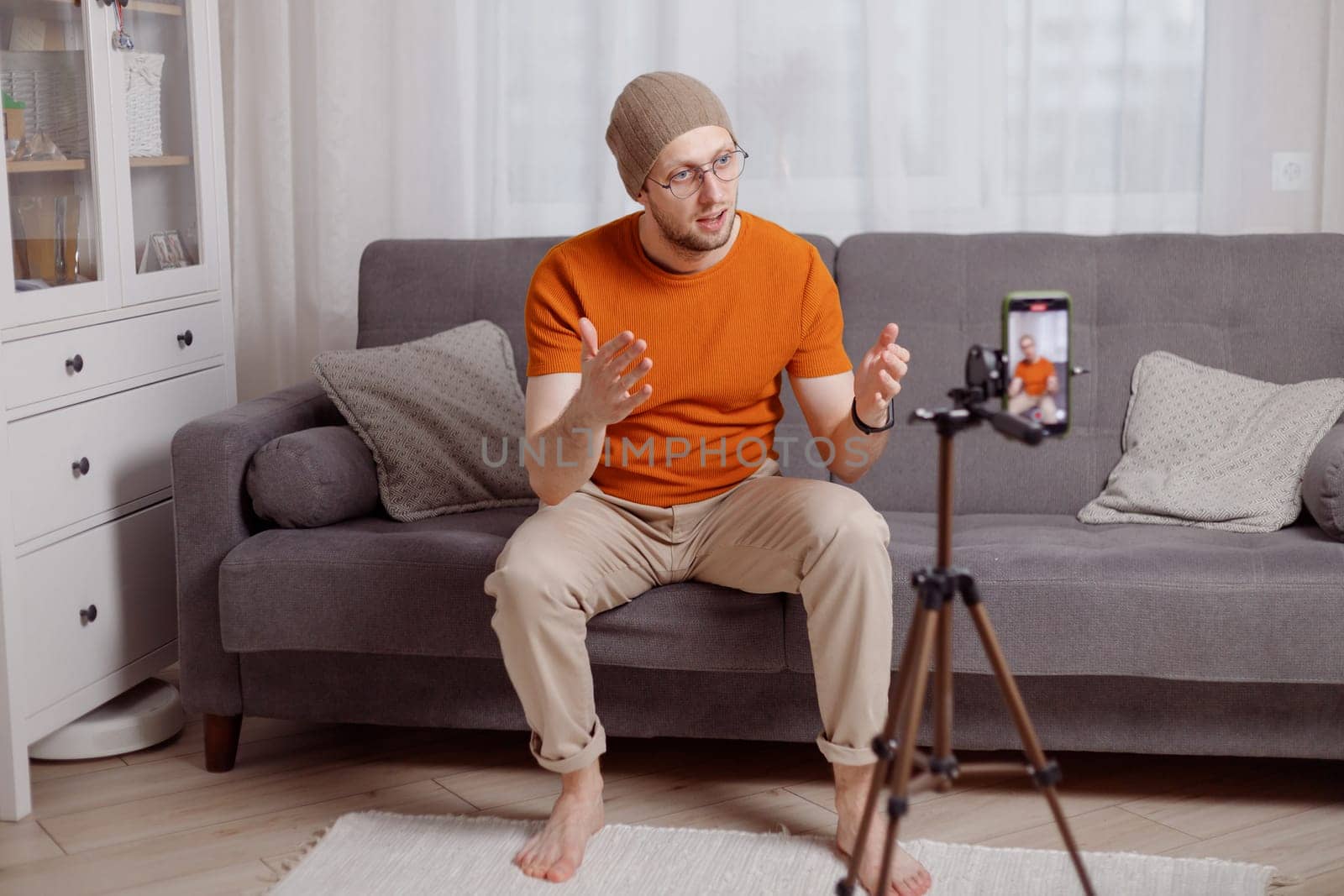 Hipster man blogger recording vlog on mobile phone sit on couch in living room and explain and gesturing with hands.