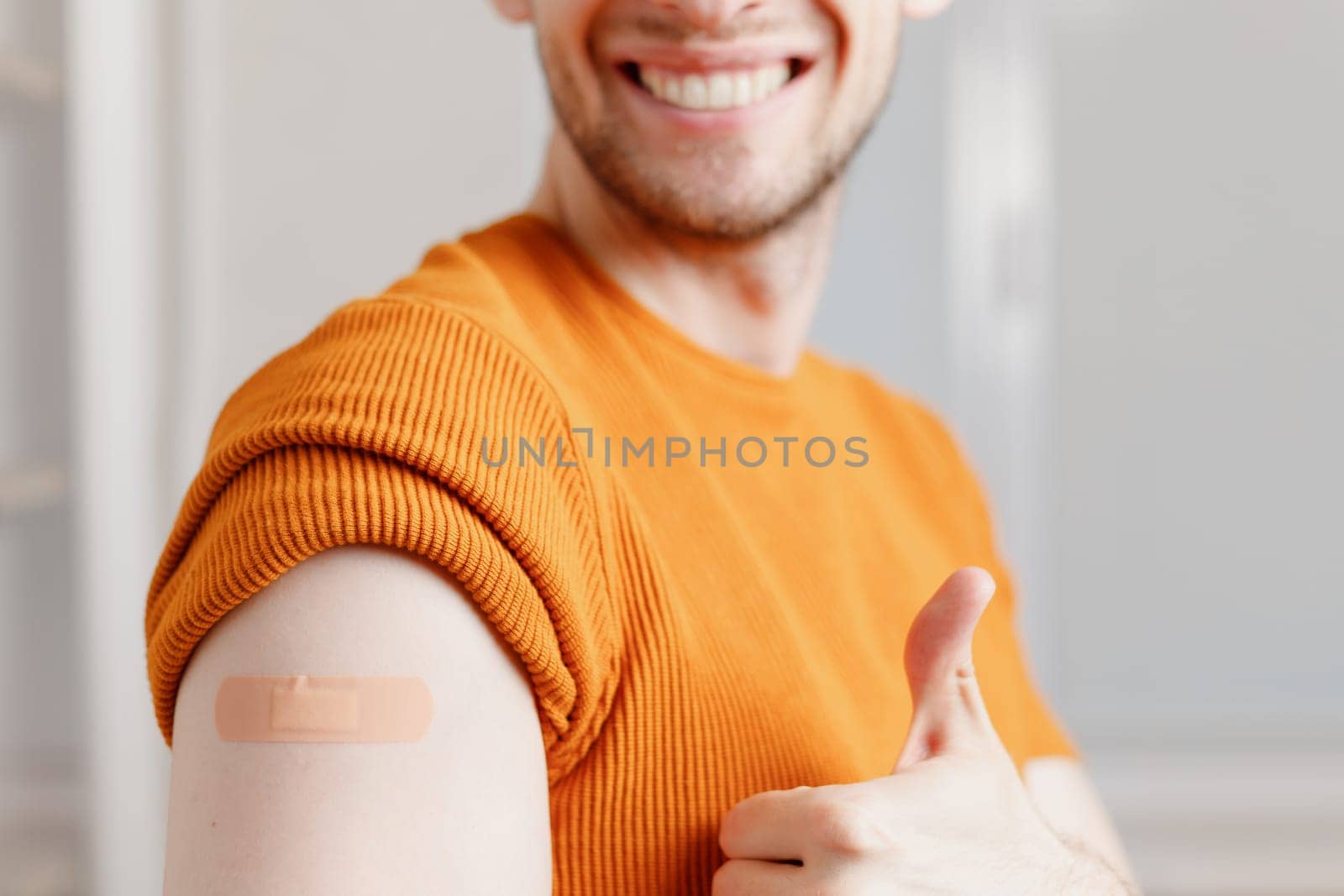 Smiling young man with adhesive bandage plaster on shoulder showing thumb up gesture after injection vaccine. Close up cropped view