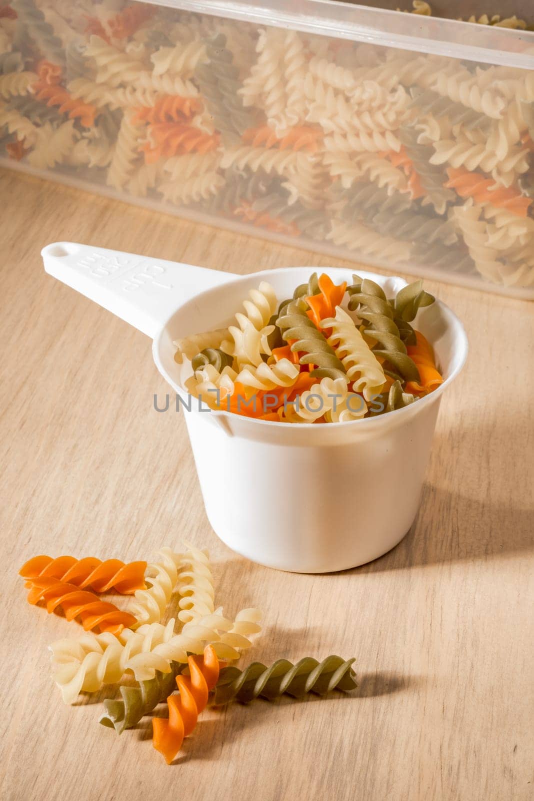 Three different colours of fussily fill a white plastic measuring cup, with a plastic container filled with more fussily in the back and some pieces of fussily lying on the wooden surface right in front.