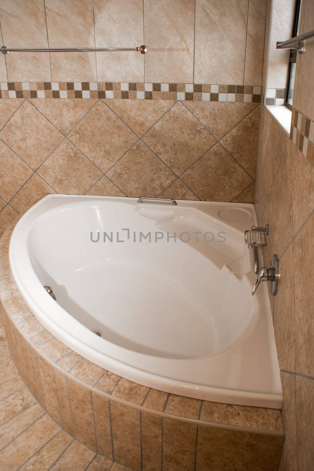 A white corner bath in a tiled bathroom, the tiles are of different shades of brown
