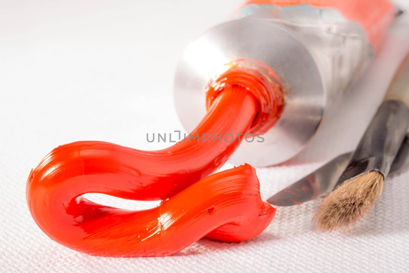 Scarlet Lake oil paint, of an orange coulour is squeezed out of the tube on to a white canvass and liles next to a used paint brush and paint knife.