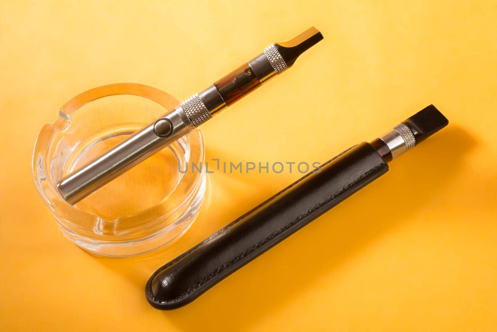 two electronic cigarettes, one in a clean glass ashtray, and the other in a black leather pouch, lie next to each other on a dark yellow background.