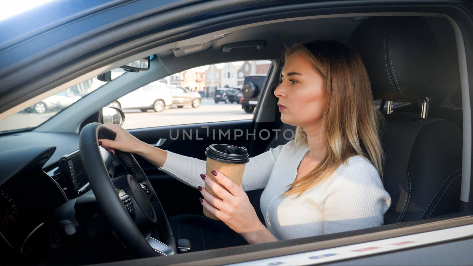 Portrait of beautiful blonde woman driving a car and drinking coffe from paper cup.