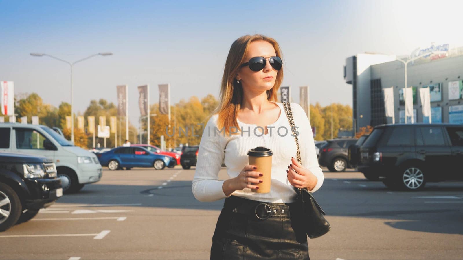 Toned portrait of young businesswoman drinking coffee from paper cup while walking on car parking to her office.