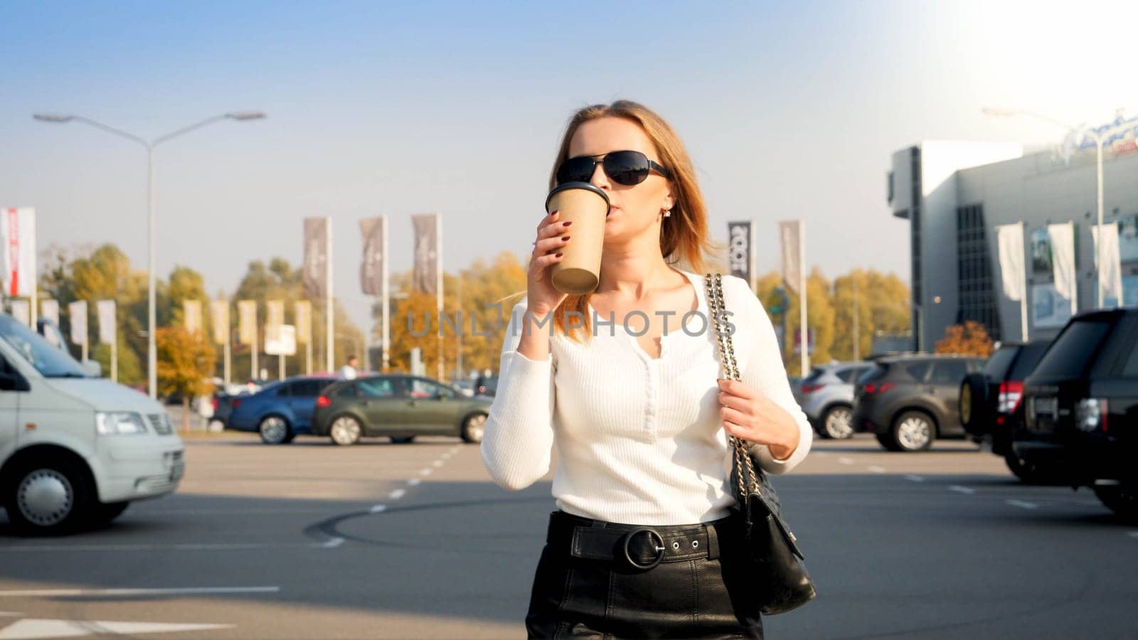 Portrait of stylish blonde woman drinking coffee on car parking at shopping mall.