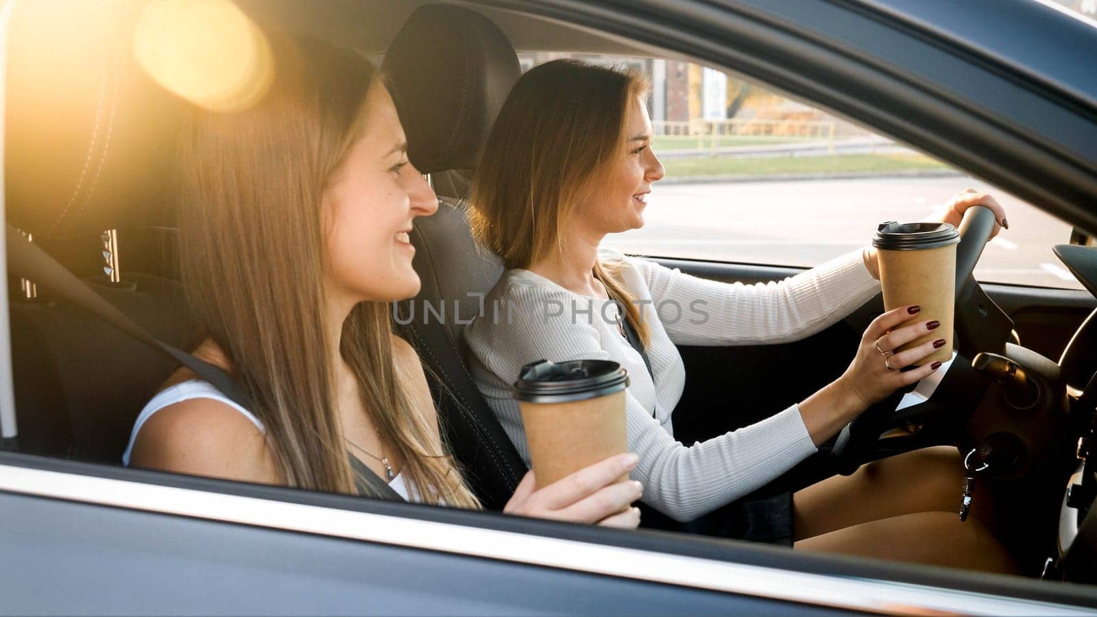 Portrait of two happy smiling girls drinking coffee while riding in car at sunset.