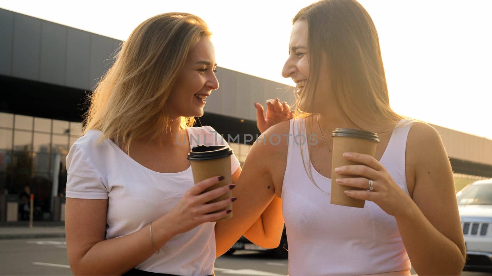 Portrait of two girl friends drinking coffe and laughing on city street at sunset.