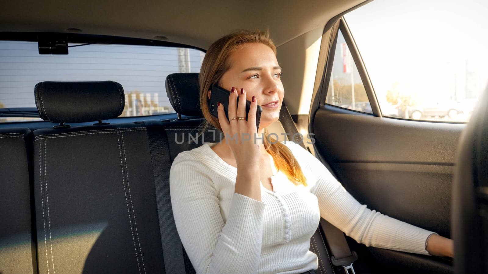 Portrait of beautiful blonde businesswoman talking by phone while riding on car back seat.