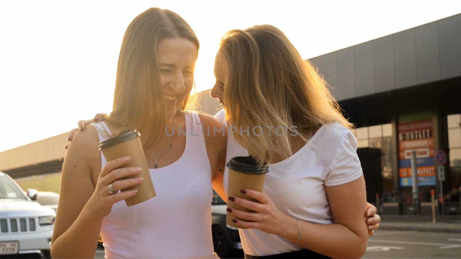 Portrait of two happy smiling girls laughing and drinking coffee while walking on city street at sunset.
