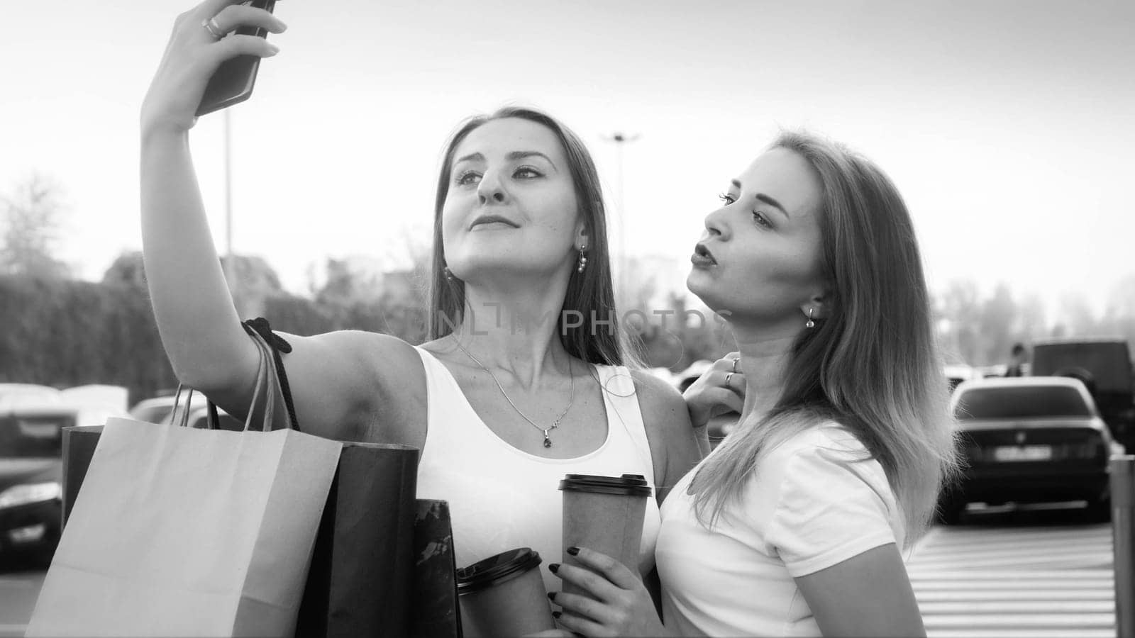 Black and white portrait of two sexy girls with shopping bags making selfie photo on shopping mall car parking.