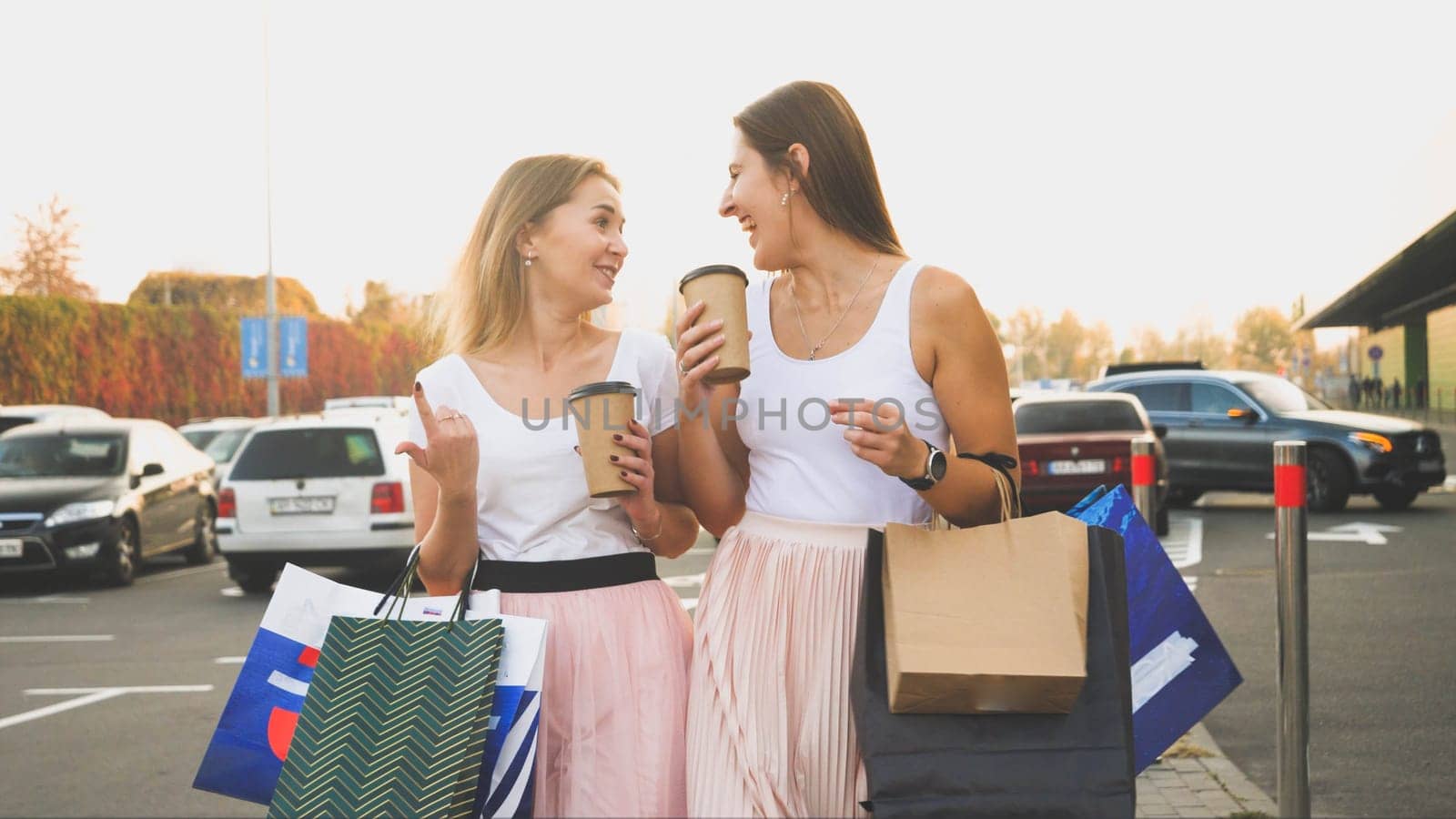 Toned photo of two women with paper bags talking and smiling while walking on car parking at shopping mall.
