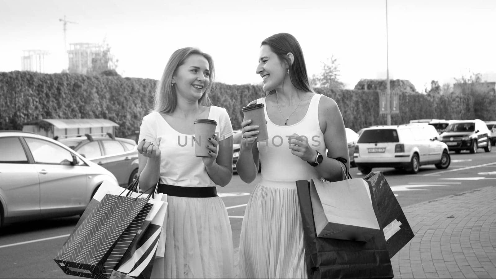 Two smiling girls with paper shopping bags talking while walking on car parking at big mall.